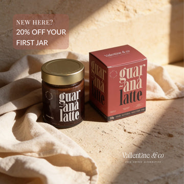 A jar of guarana latte and its matching red box are styled on beige fabric and stone with warm sunlight shadows. Text in the corner reads 'NEW HERE? 20% OFF YOUR FIRST JAR' and the brand 'Valentino & co' best coffee alternative.